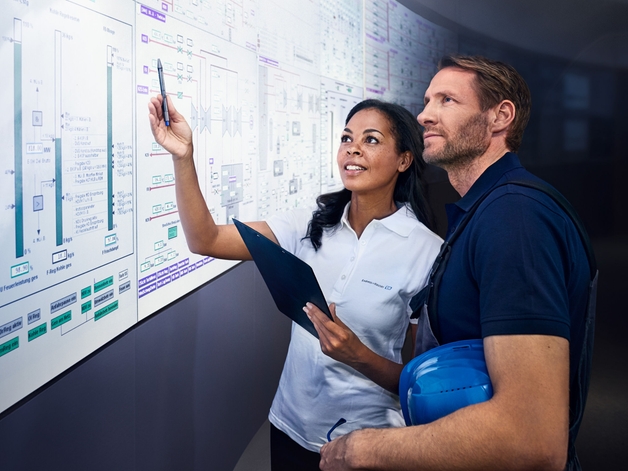 Two engineers discuss in front of a large display of an industrial control room.